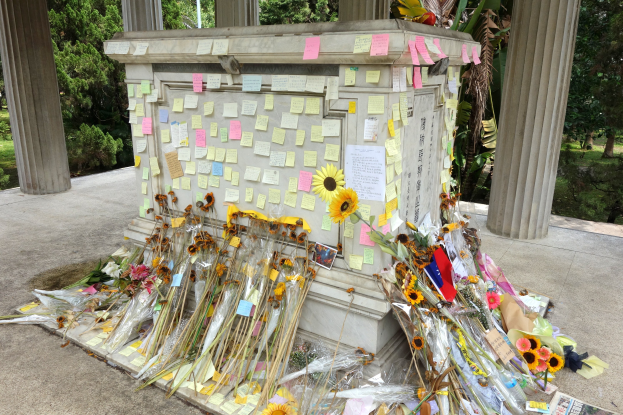Ein Gedenkstein umgeben von Bäumen, Pflanzen und Gras, geschmückt mit Post-it-Notizen und Blumen.