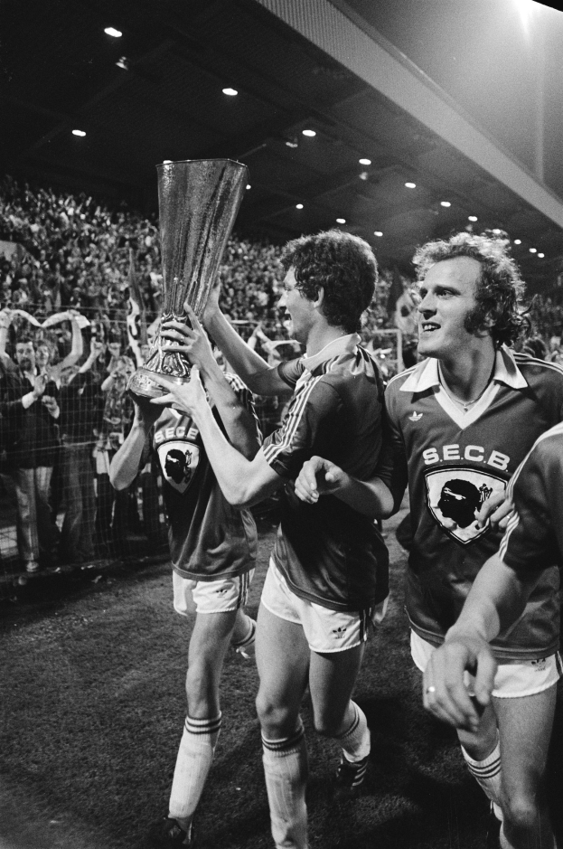 Schwarzes und weißes Foto von Fussballspielern, die einen Pokal auf einem Feld halten und ihren FA-Cup-Finalsieg feiern, mit sichtbaren Zuschauern hinter einem Zaun.