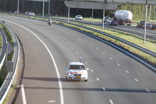 Ein Auto auf einer Autobahn mit einem stationären Polizeiwagen am Rande, andere Fahrzeuge im Verkehr und Bäume und Straßenstrukturen im Hintergrund.