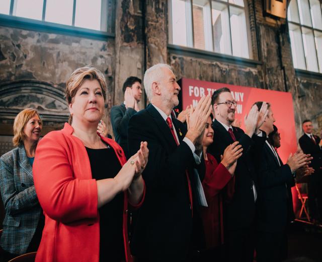 Eine Gruppe von Menschen steht vor einer Menge und klatscht in die Hände, mit einem Banner mit Text im Hintergrund.