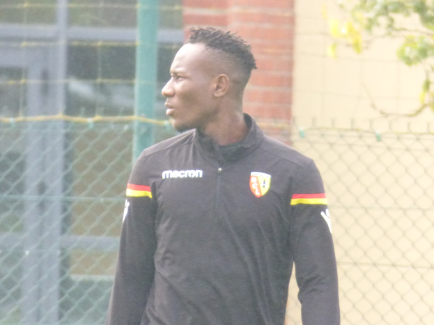 Ein Mann in einer schwarzen Jacke steht auf einem Fussballfeld, schaut nach vorne und hat einen entschlossenen Ausdruck, umgeben von einem Zaun und einem Baum auf der rechten Seite.