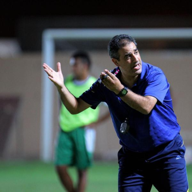 Ein Mann in einem blauen T-Shirt und Hose, der ein Mikrofon hält, steht auf einem Fußballfeld und gestikuliert, mit einer Person in einem grünen Kleid, einem Torpfosten und einer Wand im Hintergrund.