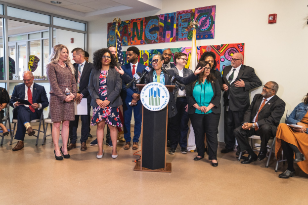 Eine Gruppe von Menschen steht in der Nähe eines Podiums mit einem Mikrofon und einer Tafel mit der Aufschrift "Sancho-Stadtrat tritt dem Stadtrat bei", mit sitzenden und stehenden Teilnehmern in einem Raum mit Flaggen, Feuerlöscher und Deckenleuchten.