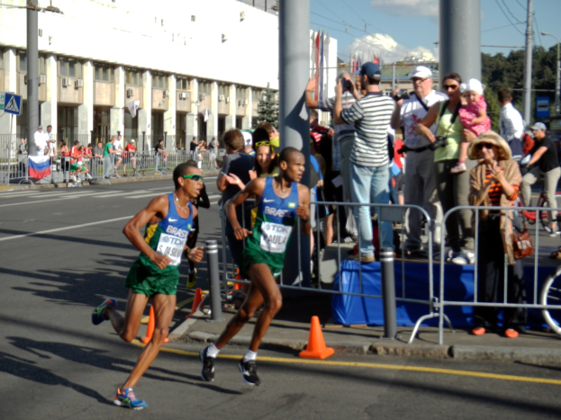 Zwei Männer bei einem Marathon auf einer Stadtstraße, umgeben von einer Zuschauermenge auf dem Bürgersteig und verschiedenen städtischen Elementen im Hintergrund.
