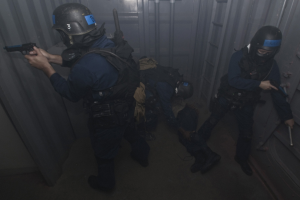 Eine Gruppe von Polizisten in Einsatzausrüstung, einer hält eine Waffe, neben einem Metallcontainer und einer Leiter in einem beengten Raum.