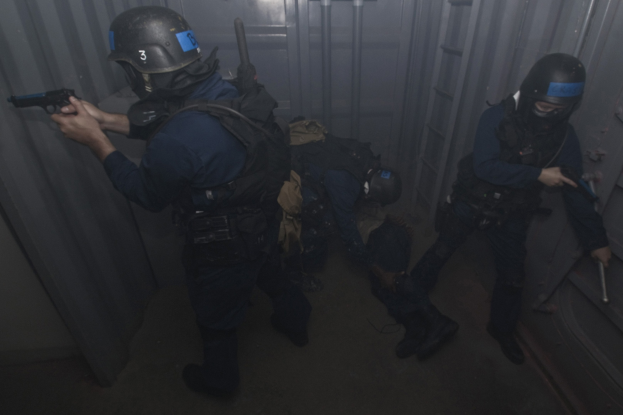 Eine Gruppe von Polizisten in Einsatzausrüstung, einer hält eine Waffe, neben einem Metallcontainer und einer Leiter in einem beengten Raum.