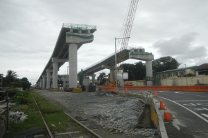 Baustelle mit einer Brücke im Hintergrund, eine Straße mit Absperrbaken auf der rechten Seite, Steine und Gras am Boden, eine Bahnschiene auf der linken Seite, Bäume und Gebäude auf beiden Seiten der Straße und ein bewölkter Himmel.