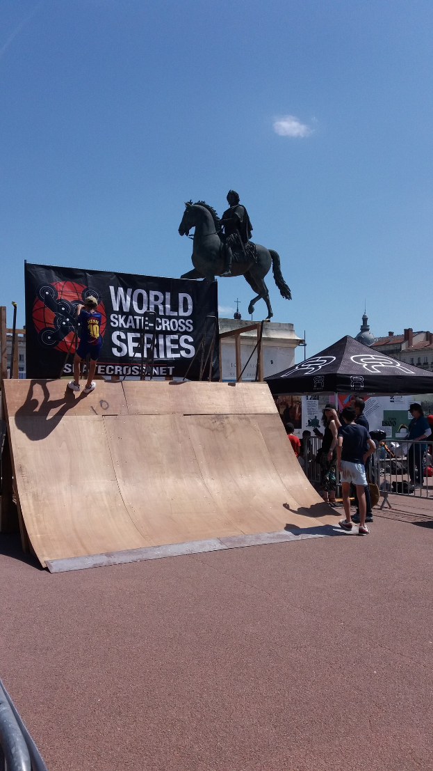 Ein Skateboarder führt einen Trick auf einer Rampe vor einem Publikum aus, mit Bannern, Gebäuden und einem bewölkten Himmel im Hintergrund.