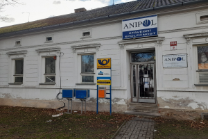 Weißes Gebäude mit einer Tafel, die "Anipol Post Office" lautet, mit Fenstern, einer Tür, einer Tafel mit Text, Pfosten, einem Weg, Gras, trockenen Blättern, einem Baum und einem bewölkten Himmel.