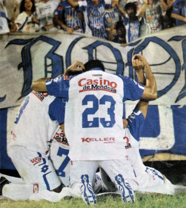 Fußballspieler feiern ein Tor auf dem Feld mit jubelnden Zuschauern und einem Banner im Hintergrund.