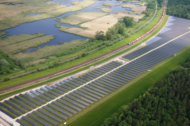 Eine Luftaufnahme einer Solar-Farm mit zahlreichen Solar-Panelen auf einem Feld, umgeben von Bäumen, Gras und Wasser, mit einem Zug, der auf einem nahen Bahngleis fährt.