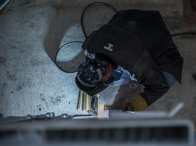 Ein Schweißer in Schutzausrüstung nutzt eine Schweißmaschine an einem Metallstück, mit Werkzeugen und Gegenständen, die auf dem Boden verstreut sind.