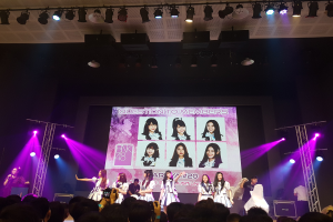 Eine Gruppe von Menschen performs auf einer hell erleuchteten Bühne mit Trägern, während eine Menge darunter das Ereignis aufzeichnet; ein großer Bildschirm im Hintergrund zeigt Bilder der K-Pop-Gruppe BK48.