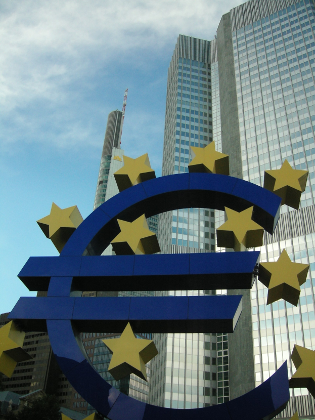 Euro-Zeichen vor dem Hauptsitz der Europäischen Zentralbank in Frankfurt, Deutschland, mit dem Gebäude umgeben von Bäumen und einem bewölkten Himmel.