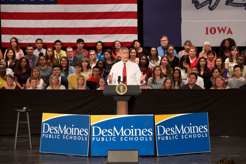 Ein Mann in einem weißen Hemd und roter Krawatte steht an einem Podium mit drei Texttafeln, einem Mikrofonständer und einem sitzenden Publikum im Hintergrund, mit einer Flagge und zusätzlichem Text.
