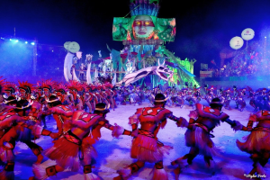 Eine Gruppe von Menschen in bunten Kostümen tanzt auf einer Bühne vor einem sitzenden Publikum, mit Lichtern, Stangen, Ballons und einem Bildschirm, der eine Person in den Hintergrund zeigt, unter einem sichtbaren Himmel, bei der Rio-Karneval 2014.