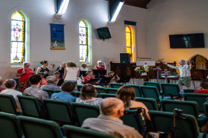 Eine Gruppe von Menschen sitzt auf Stühlen in einer Kirche, ein Mann steht vorne mit einem Mikrofon, umgeben von Musikinstrumenten, einem Tisch mit einer Vase, einem Lautsprecher, einem Banner an der Wand, Fenstern, einem Fernseher und Deckenleuchten.