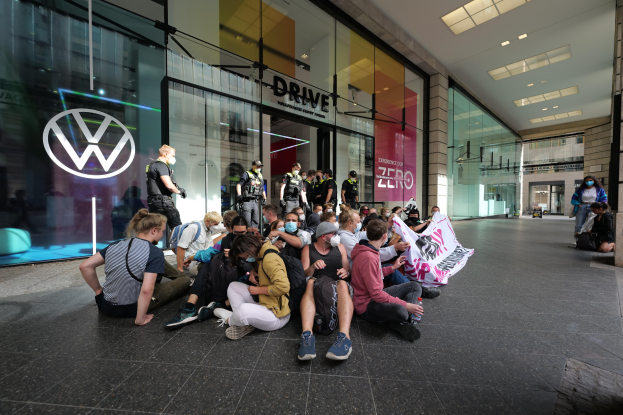 Eine Gruppe von Menschen sitzt vor einer Volkswagen-Vertretung mit Glaswänden auf dem Boden und hält eine Fahne mit Text, unter Deckenleuchten.