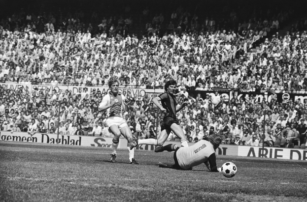 Schwarzes Foto von Männern, die auf einem Feld Fußball spielen, mit Zuschauern im Hintergrund und einer 'Bundesliga 1978-79 Ajax 1-1 Hannover 96' Fahne.