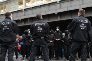 Eine Gruppe uniformierter Polizisten steht vor einer Menge von Menschen in schwarzen Uniformen und Masken mit einer Brücke und einem Gebäude im Hintergrund während einer Protestaktion in einer Stadt.
