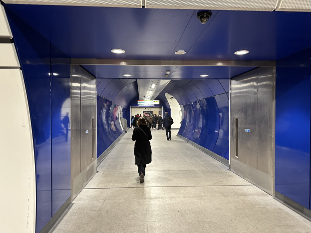 Eine Gruppe von Menschen, die durch einen blauen und weißen Tunnel mit Türen auf beiden Seiten und Lampen oben gehen, mit einer Person in Laufschuhen in der Mitte.