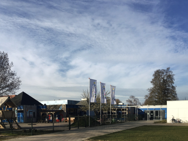 Großes Gebäude mit Metallzaun, Rasenfläche, Weg, Bäumen, Pfosten, Bannern, einem geparkten Fahrrad, nahen Häusern und einem bewölkten Himmel, als neuer Spielplatz für Kinder an der Schule identifiziert.