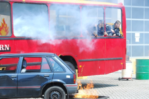 Roter Doppeldeckerbus mit Rauch aus dem Auspuff, daneben geparktes Auto, drei sichtbare Passagiere im Bus und im Hintergrund ein Gebäude mit Glasfenstern und einem Fass.