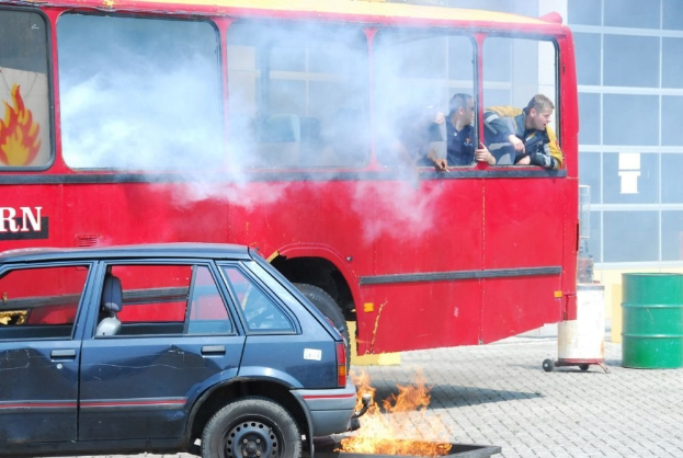 Roter Doppeldeckerbus mit Rauch aus dem Auspuff, daneben geparktes Auto, drei sichtbare Passagiere im Bus und im Hintergrund ein Gebäude mit Glasfenstern und einem Fass.