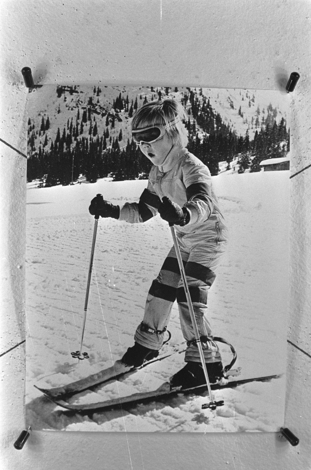 Ein junger Junge, der einen verschneiten Hügel mit Skiern, Helm, Handschuhen und Schuhen hinunterfährt und Ski-Stöcke hält, mit Bäumen und einem klaren Himmel im Hintergrund.