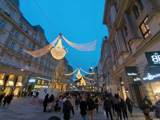 Eine belebte Stadtstraße mit Menschen, die gehen, geschmückt mit festlichen Weihnachtslichtern, die von oben hängen, flankiert von Gebäuden mit Fenstern und einem klaren blauen Himmel im Hintergrund.