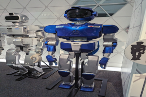 Zwei humanoide Roboter stehen nebeneinander in einem Ausstellungsraum mit einer Tafel auf der rechten Seite und einer Wand im Hintergrund.