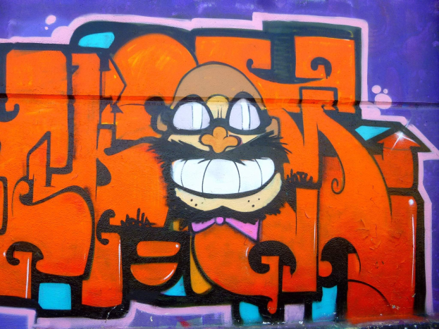 Graffiti-Gemälde eines Cartoon-Charakters mit großen Augen, einem breiten Grinsen und einem verschmitzten Gesichtsausdruck an der Seite eines Gebäudes, mit leuchtenden Farben und auffälligem Begleittext.