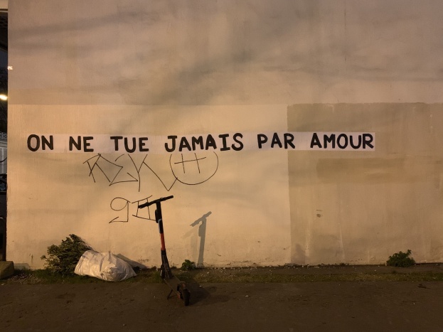 Ein Roller steht vor einer Wand mit der Graffiti-Aufschrift "On ne tue jamais par amour", mit Pflanzen und einer Abdeckung auf dem Boden und einem Gebäude, Laternenmast und Himmel im Hintergrund.