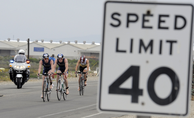 Gruppe von Radfahrern in Helmen, die eine Straße mit einem Geschwindigkeitsbegrenzungsschild entlangfahren, mit Gebäuden und Bäumen im Hintergrund unter einem klaren blauen Himmel.