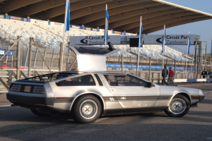 Ein silberner DeLorean DMC-12 Wagen auf einer Straße umgeben von Menschen, Flaggen, Schildern, Geländern, Stühlen und einem Schuppen, mit Bäumen und einem klaren blauen Himmel im Hintergrund.