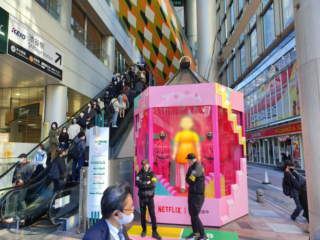 Eine Gruppe von Menschen steht vor einem Gebäude mit Glaswänden, Säulen und Texttafeln, einige tragen Masken, während hinter einem pinken Kasten mit Schaufensterpuppen und der Aufschrift "Netflix's new Netflix Booth in Tokyo" eine Rolltreppe mit Menschen dahinter zu sehen ist.