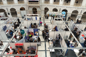 Große Besucherzahl auf einer internationalen Fachmesse mit Ständen, Bannern, Tischen, Stühlen und Luftballons in einem Raum mit Bogengängen und Säulen.