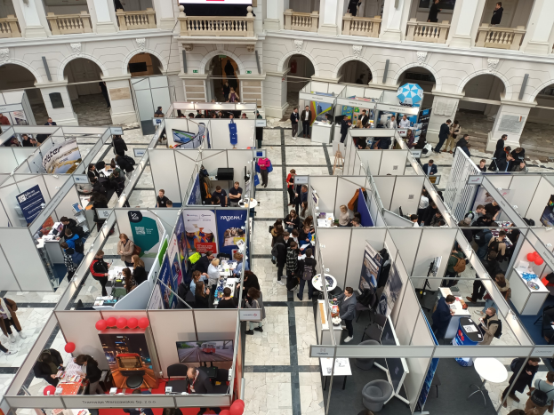 Große Besucherzahl auf einer internationalen Fachmesse mit Ständen, Bannern, Tischen, Stühlen und Luftballons in einem Raum mit Bogengängen und Säulen.