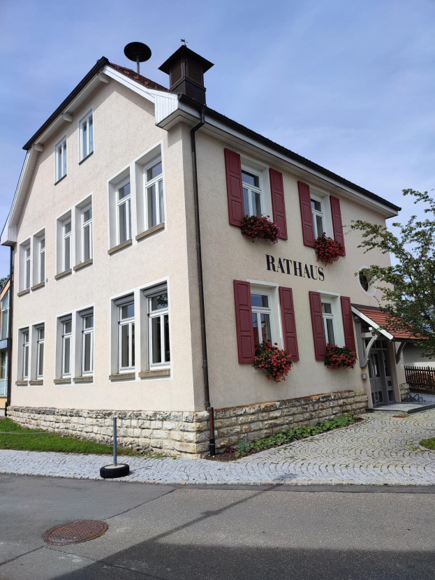Weißes Gebäude mit roten Fensterläden, umgeben von Grün und Blumen, mit einer Straße unten und Himmel im Hintergrund, als Rathaus-Hotel in Baden-Württemberg identifiziert.