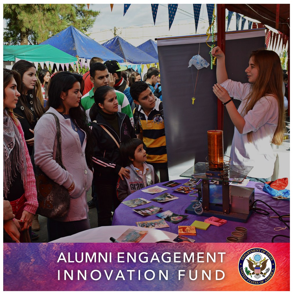 Gruppe von Menschen um einen Tisch versammelt, auf dem ein Schild mit der Aufschrift "Alumni Engagement Innovation Fund" liegt, mit Gegenständen auf dem Tisch, Zelten und Bäumen im Hintergrund unter einem klaren blauen Himmel und Text mit einem Logo unten.