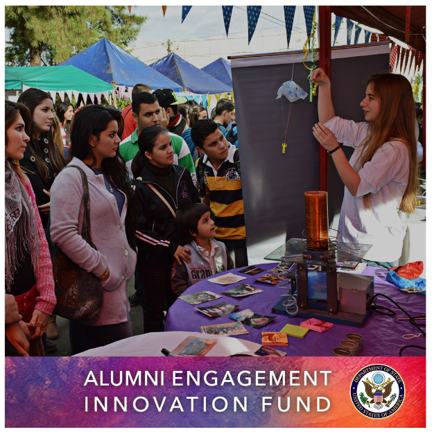 Gruppe von Menschen um einen Tisch versammelt, auf dem ein Schild mit der Aufschrift "Alumni Engagement Innovation Fund" liegt, mit Gegenständen auf dem Tisch, Zelten und Bäumen im Hintergrund unter einem klaren blauen Himmel und Text mit einem Logo unten.
