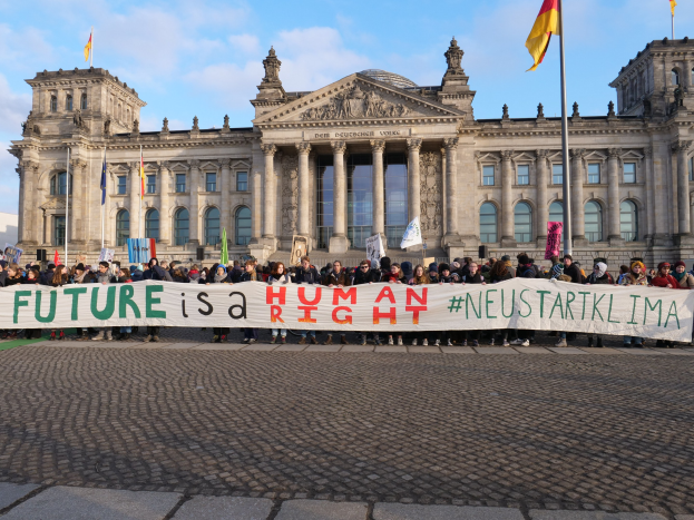 Eine Gruppe von Menschen hält ein Transparent mit der Aufschrift "Zukunft ist ein Mensch" vor dem Reichstaggebäude in Berlin, wobei die Flagge des Deutschen Bundestages zu sehen ist.