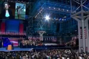 Eine große Zuschauermenge sitzt einer Bühne mit einem Rednerpult, einem Mikrofon, Flaggen, einem Bildschirm und Lichtausrüstung zugewandt, was an eine Präsidentschaftsdebatte erinnert.