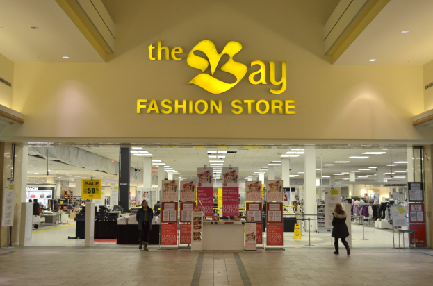 Außenansicht eines Bay Fashion Stores in einem Einkaufszentrum mit Menschen, die gehen, Texttafeln an den Wänden, Säulen, Deckenleuchten und Hintergrundanzeigen.