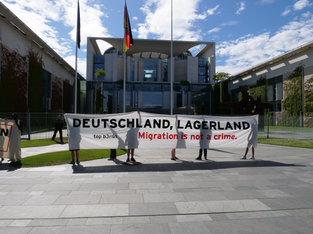 Eine Gruppe von Menschen mit einem Transparent mit der Aufschrift "Deutschland, Lagerland Migration ist kein Verbrechen" vor einem umzäunten Gebäude mit Bäumen und Fahnen.