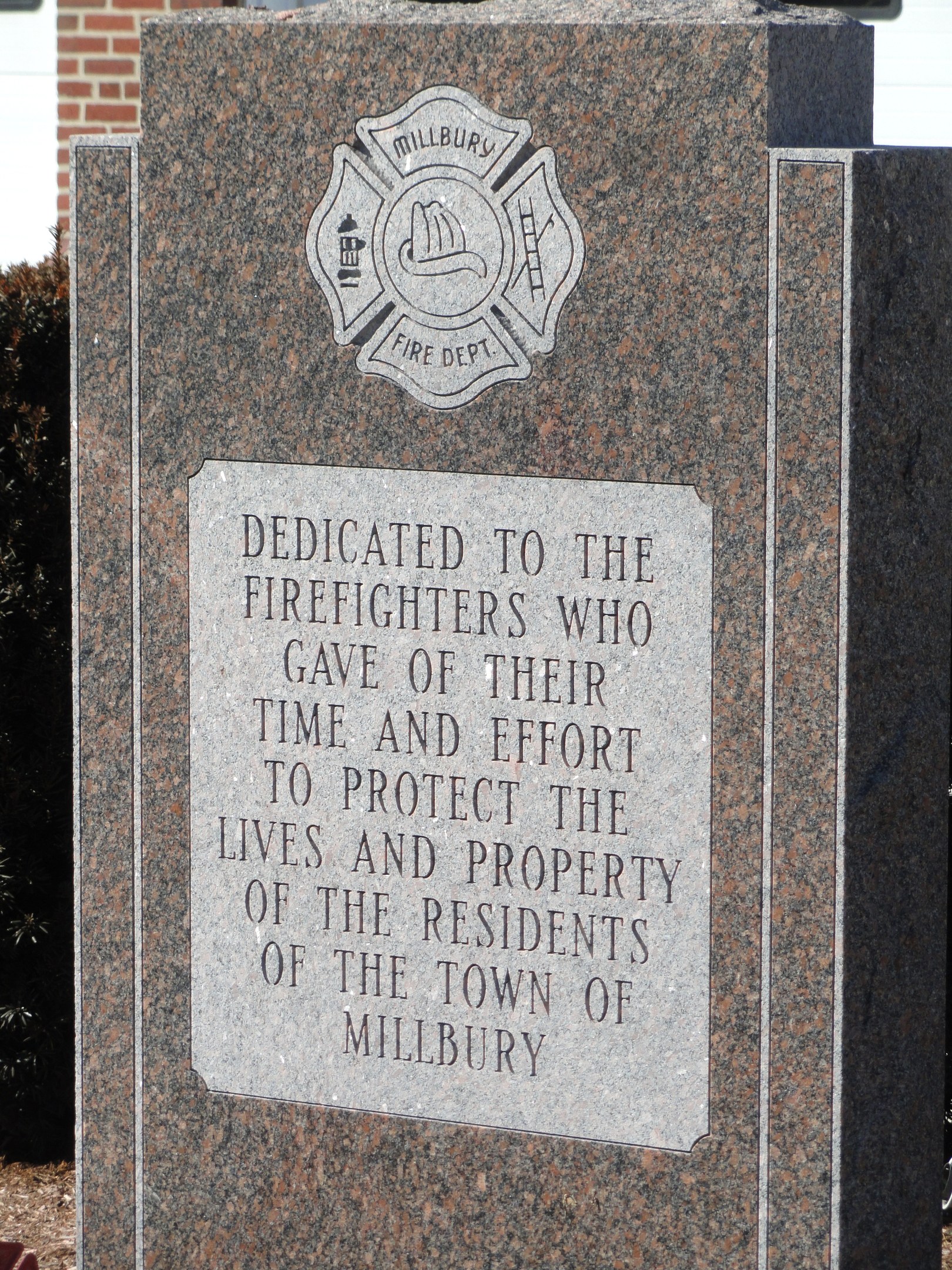 Denkmalstein mit einer Gedenktafel für Feuerwehrleute in Millbury, vor einem Backsteingebäude mit umliegender Vegetation.