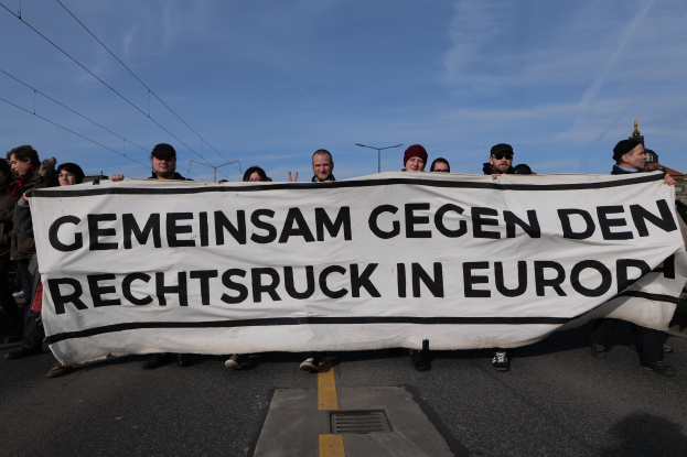 Eine Gruppe von Menschen steht auf der Straße und hält ein Banner mit der Aufschrift "Gemeinsam Gegen den Rechtsruck in Europa" mit Straßenlaternen, Strommasten, Kabeln, Gebäuden und einem bewölkten Himmel im Hintergrund.