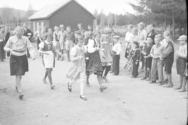 Schwarzes Foto von Kindern, die Fußball spielen, mit einer Menge, die Fahnen hält, im Hintergrund Bäume und ein Haus.
