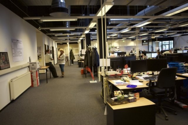 Großes Großraumbüro mit Menschen an Schreibtischen, Computern, Monitoren, Stühlen, Papieren, Büchern, Kartons, an der Wand hängenden Kleidung, Wandtafeln mit Text und Bildern, Deckenleuchten und Glasfenstern im Hintergrund.