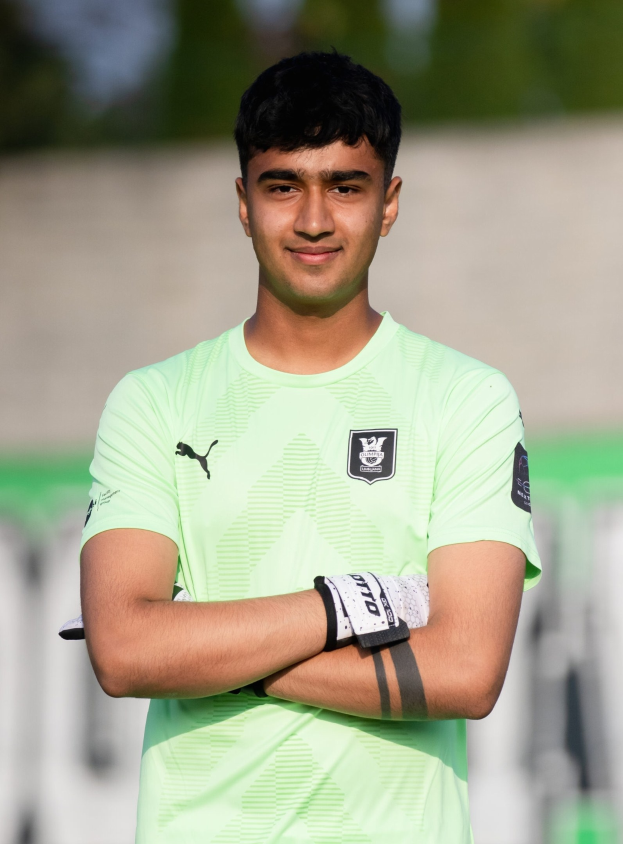 Ein junger Mann in einem grünen T-Shirt, wahrscheinlich ein Torwart, steht auf einem Fußballfeld und hat die Arme verschränkt, während er lächelt, und der Hintergrund ist unscharf.